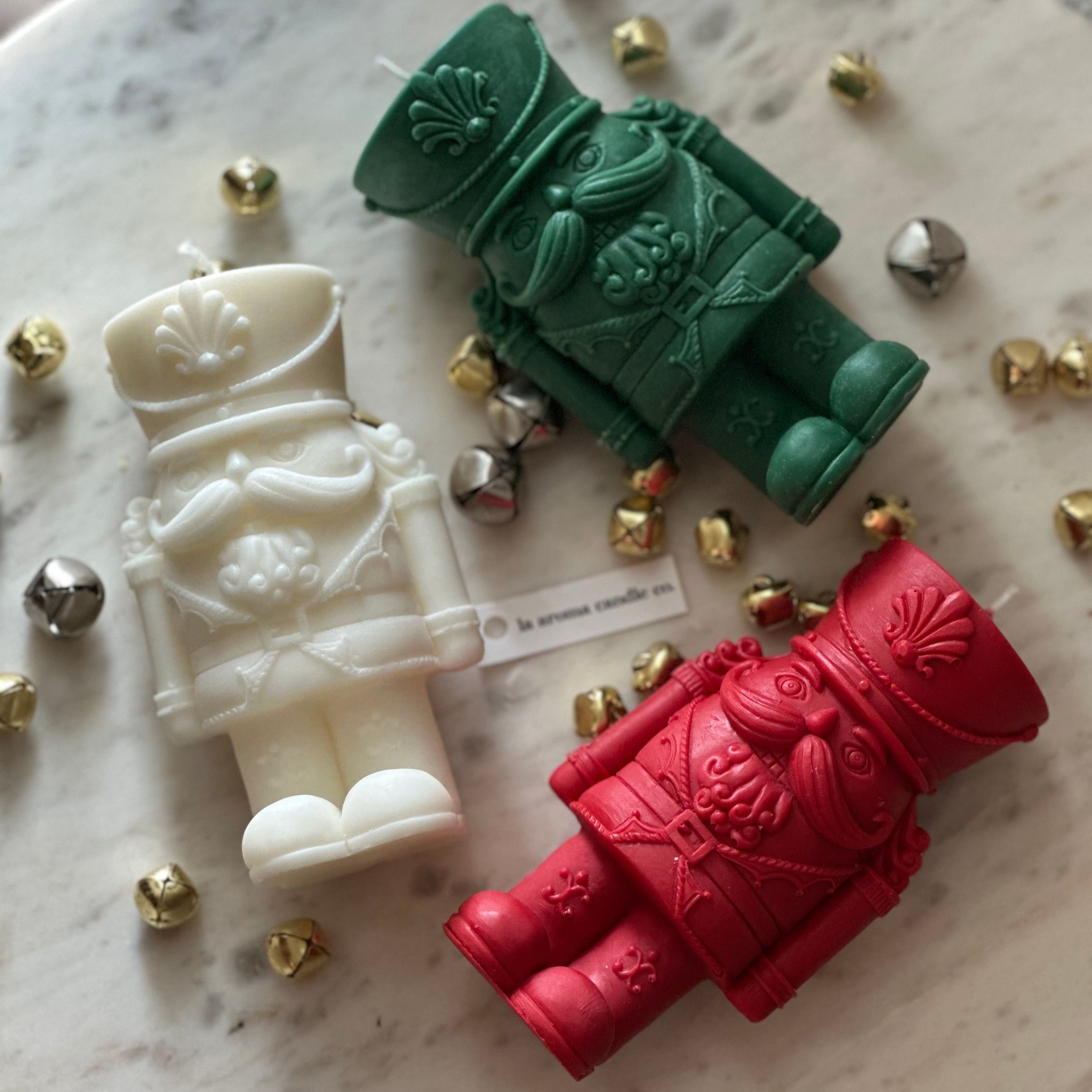 Three toy soldier-shaped candles in white, green, and red on a marble surface with small metallic objects.