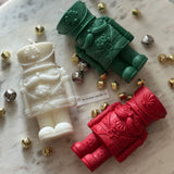 Three toy soldier-shaped candles in white, green, and red on a marble surface with small metallic objects.